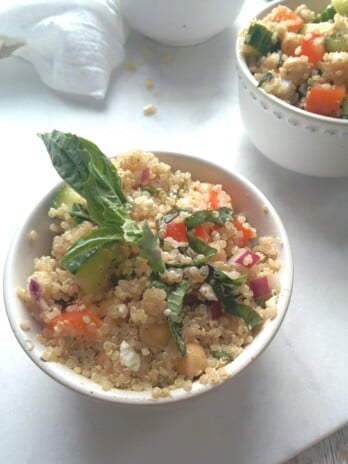 Mediterranean and Chickpea Quinoa Salad in a white round bowl. Ingredients include quinoa, chicken stock, red wine vinegar, grape tomatoes, onion, chickpeas, feta, basil, parsley.