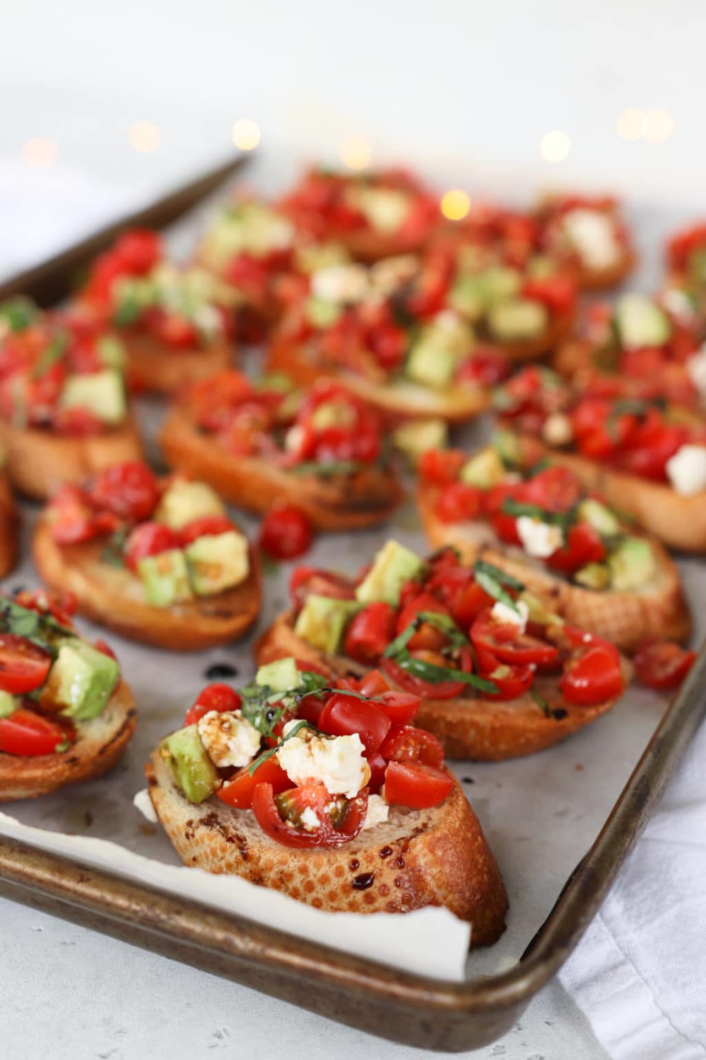 tomato and avocado bruschetta line a parchment paper lined baking sheet. One bruschetta topped crostini is in focus in the front while the other pieces fade into the background