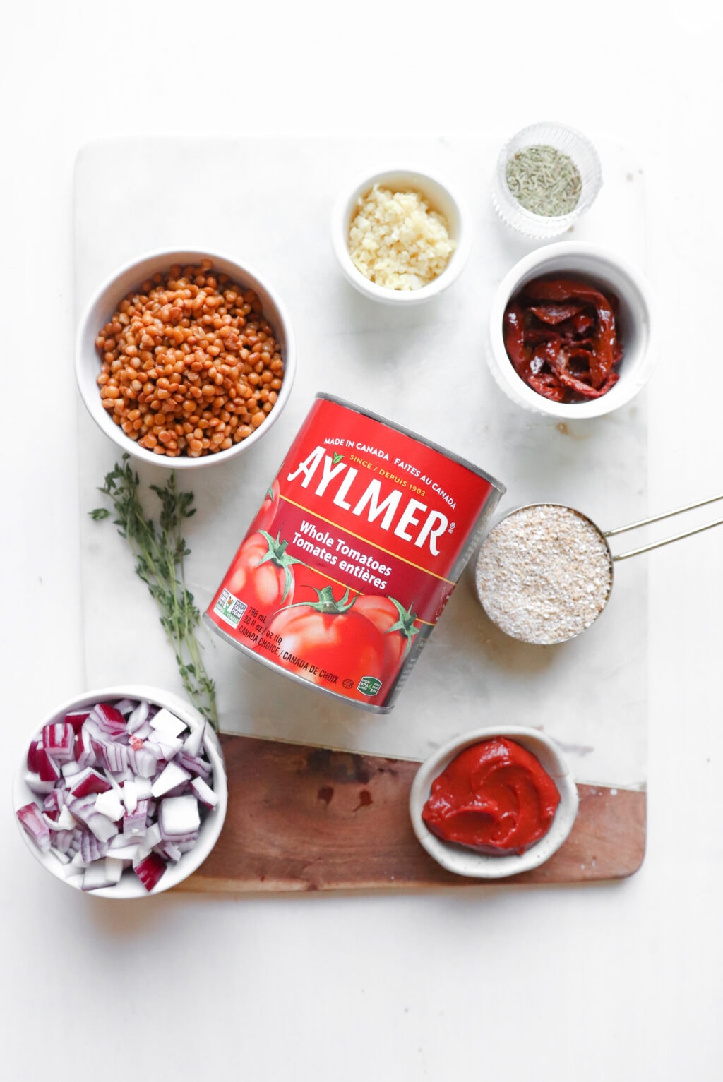 Ingredients of roasted tomato lentil soup are laid out in bowls. Moving left to right are red lentils, garlic, sundried tomatoes, steel cut oats, red onion, thyme, canned tomatoes, steel-cut oats, and tomato paste