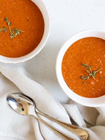two bowls of red tomato soup next to two spoons. The soup is topped with thyme