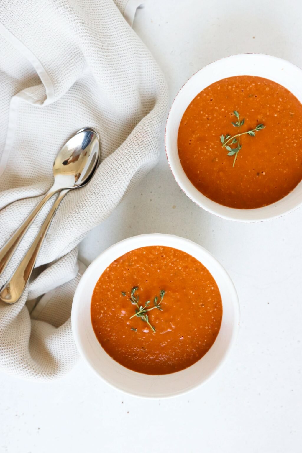 two bowls of red tomato soup next to two spoons. The soup is topped with thyme
