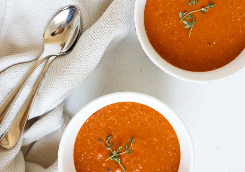 two bowls of red tomato soup next to two spoons. The soup is topped with thyme