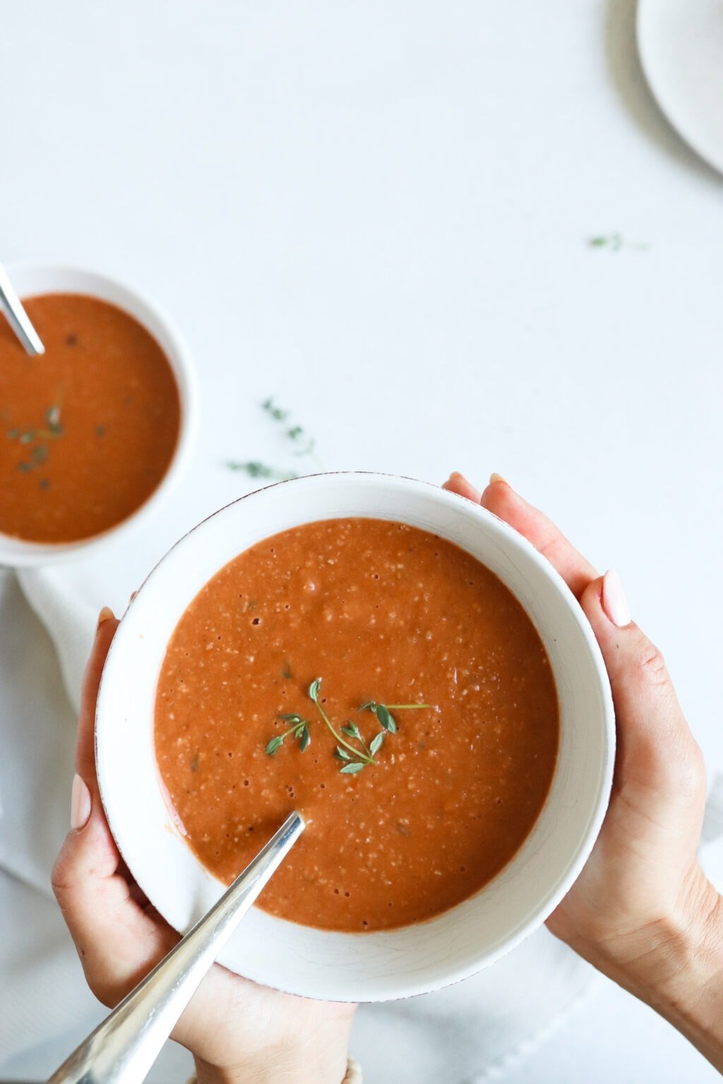 two hands are holding a bowl of roasted tomato soup towards the camera. The soup is in a white bowl and is topped with sprigs of fresh thyme