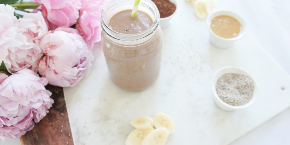 Creamy Chocolate Banana Chia Smoothie in a mason jar with a white and green striped straw on a marble food photography board. Ingredients include almond milk, chia seeds, cocoa, Greek yogurt, banana, nut butter, dates.