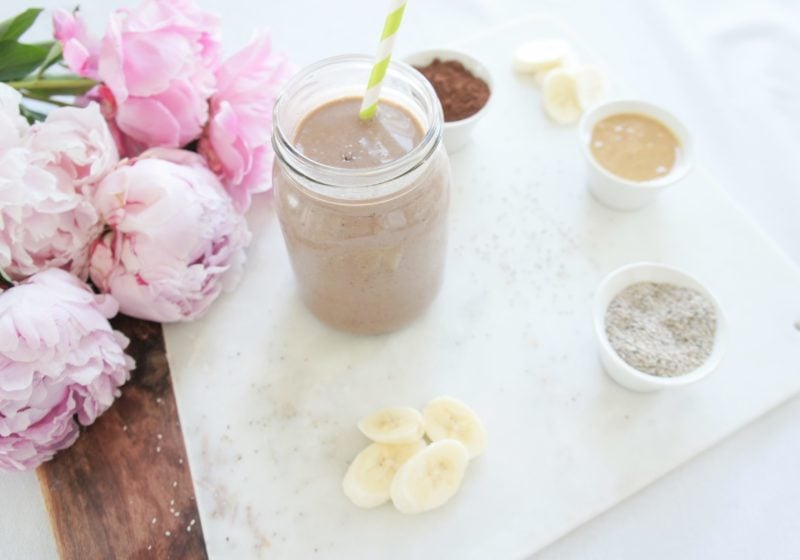Creamy Chocolate Banana Chia Smoothie in a mason jar with a white and green striped straw on a marble food photography board. Ingredients include almond milk, chia seeds, cocoa, Greek yogurt, banana, nut butter, dates.