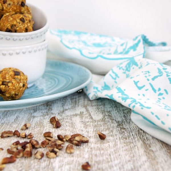 Pumpkin Pie Energy Bites placed in a stack of white round bowls on a blue plate. Ingredients include pumpkin puree, quick oats, dark chocolate chips, cinnamon, pecans, dates.