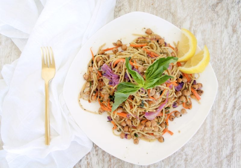 Lemon Pesto Rainbow Chickpea Pasta served in a white bowl with a gold fork and white kitchen towel placed beside it. Ingredients include spaghetti noodles, zucchini, purple cabbage, garlic, chickpeas.