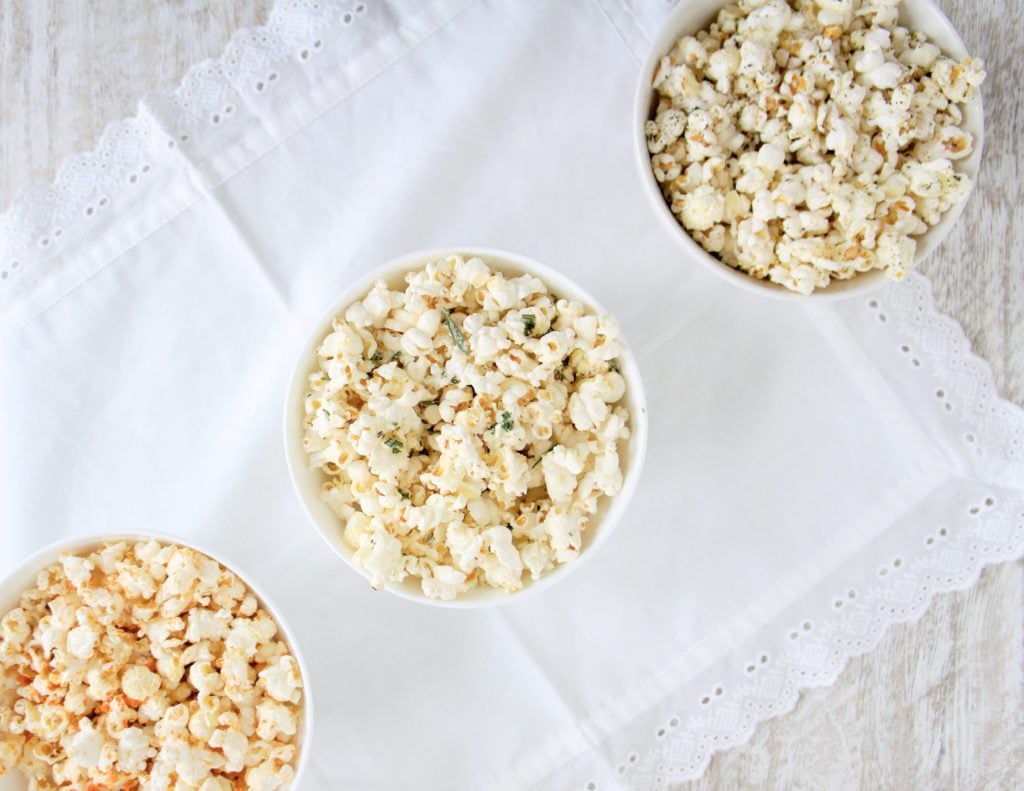 Popcorn three different ways served in white round bowls placed on a white table cloth.