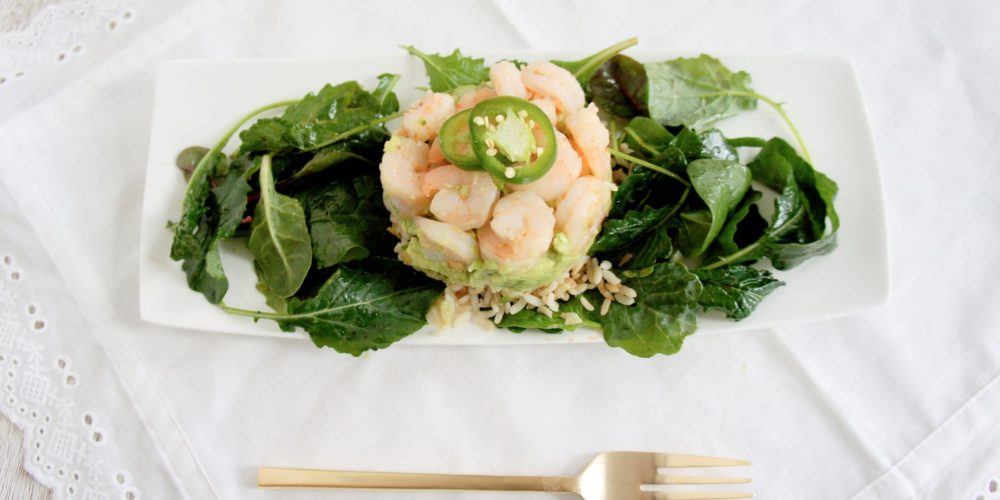 Brown Rice and Avocado Shrimp Stack with Tangy Asian Kale Salad on a white serving dish. Ingredients include brown rice, avocado, jalapeno, avocado oil, garlic, shrimp, baby kale.