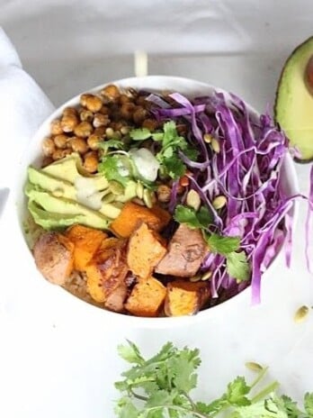 Buddha Bowl with Lemon Tahini Dressing served in a white round bowl. Ingredients include brown rice, yam, chickpeas, avocado oil, cabbage, carrots, avocado, pumpkin seeds, cilantro.