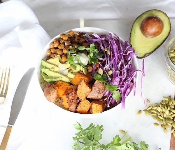 Buddha Bowl with Lemon Tahini Dressing served in a white round bowl. Ingredients include brown rice, yam, chickpeas, avocado oil, cabbage, carrots, avocado, pumpkin seeds, cilantro.
