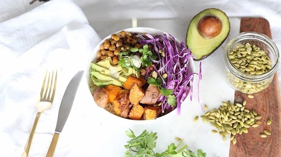 Buddha Bowl with Lemon Tahini Dressing served in a white round bowl. Ingredients include brown rice, yam, chickpeas, avocado oil, cabbage, carrots, avocado, pumpkin seeds, cilantro.