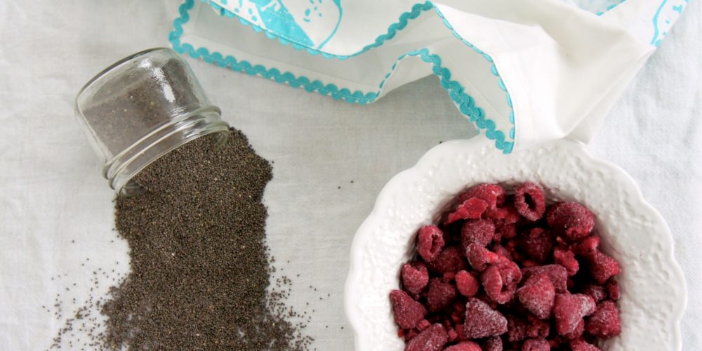 A jar of chia seeds tipped over beside a round white bowl of raspberries.