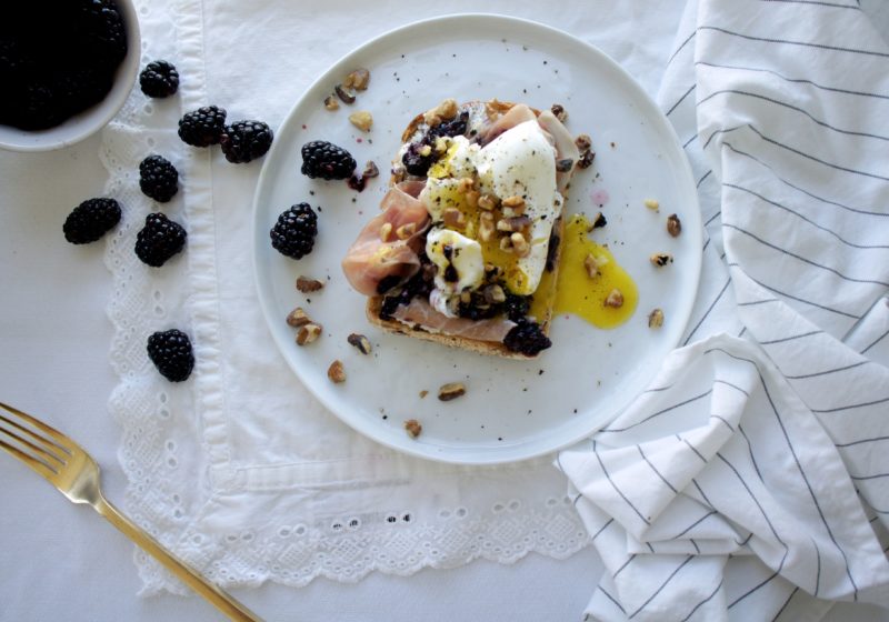 Blackberry Ricotta Toast with Poached Egg and Toasted Walnuts on a white round plate. Ingredients include vinegar, eggs, sprouted grain toast, ricotta cheese, blackberries, prosciutto, walnuts, cracked black pepper.