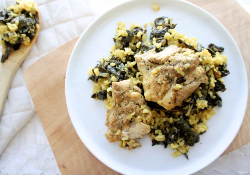 One Pan Turmeric Lemon Rice and Chicken with Kale served on a white round plate placed on a wooden cutting board. Ingredients include chicken thighs, pepper, olive oil, shallot, cumin, turmeric, curly kale, brown rice, lemon, chicken stock, Greek yogurt.