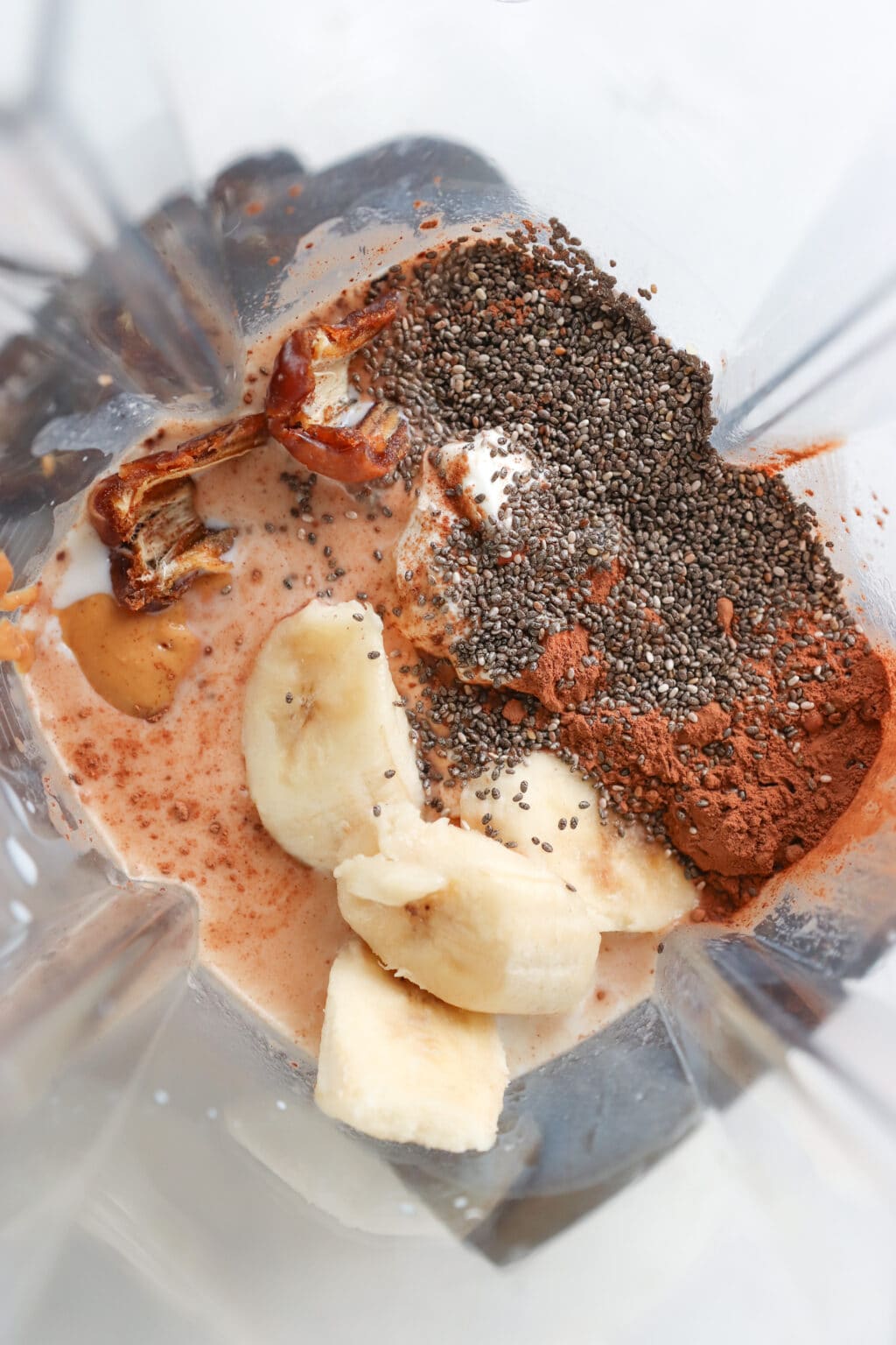 Ingredients for a 5 minute chocolate banana chia seed smoothie in a blender, including milk, chia seeds, cocoa, Greek yogurt, banana, nut, and a medjool date