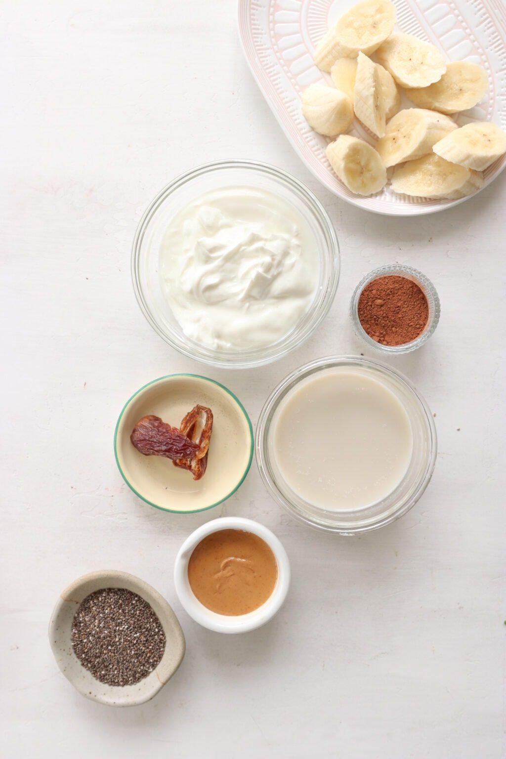 Ingredients for a 5 minute chocolate banana chia seed smoothie in glass bowls, including milk, chia seeds, cocoa, Greek yogurt, banana, nut, and a medjool date