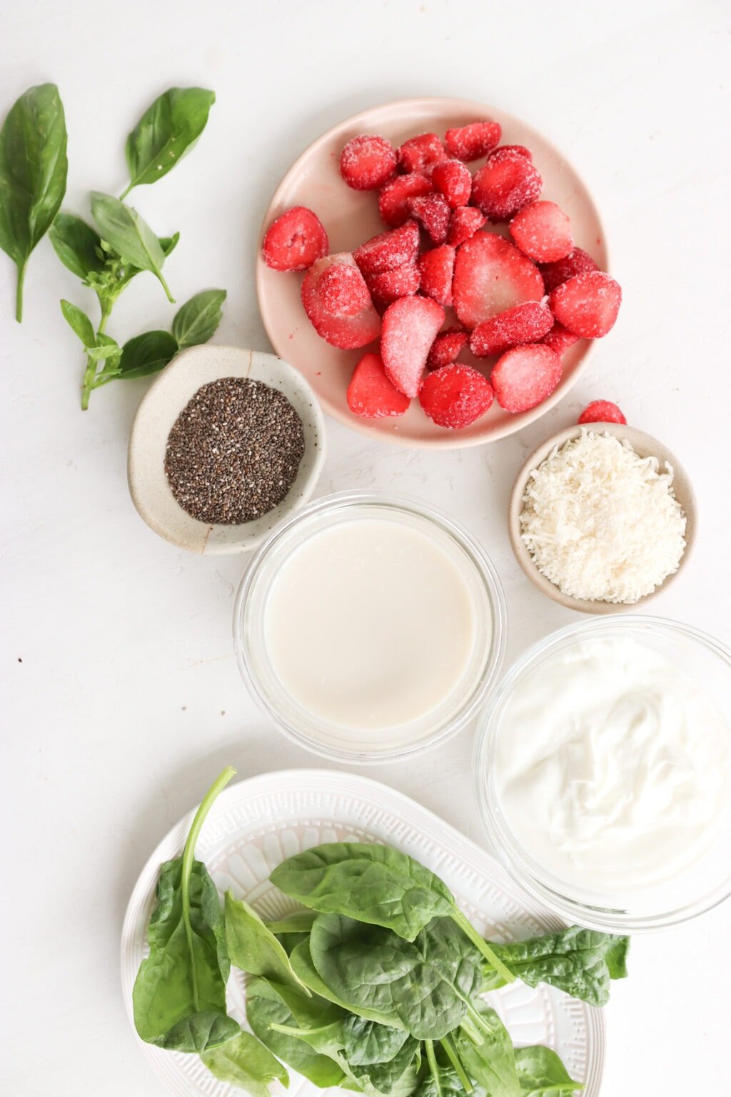 Ingredients for 5 Minute Frozen Strawberry & Basil Smoothie in glass bowls on a marble counter, including milk, Greek Yogurt, chia seeds, coconut flakes, strawberries, basil, and spinach