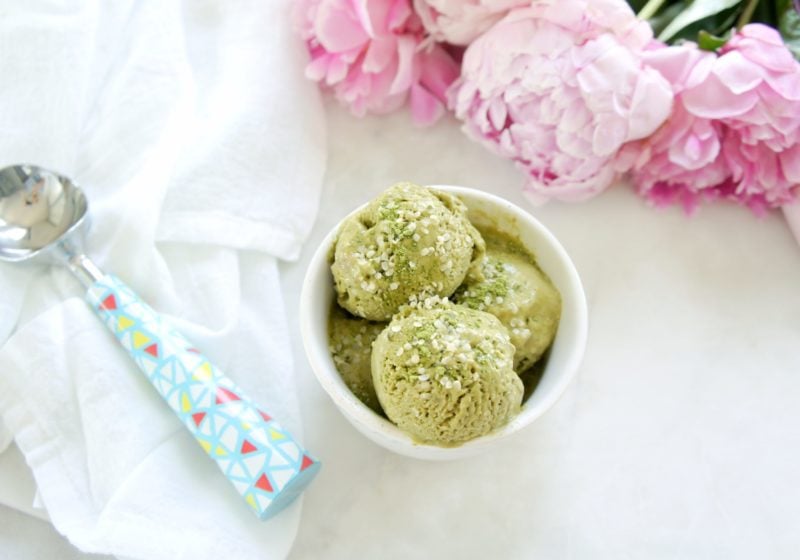 Matcha Banana Hemp Ice Cream scooped into a white round bowl placed on a white surface with an ice-cream scoop, white kitchen towel, and pink peonies beside it.