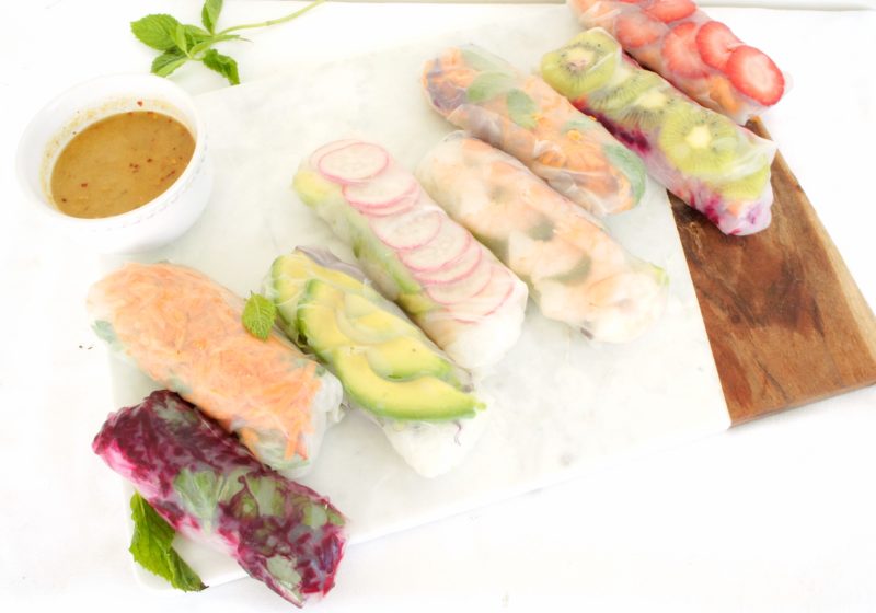 Summer Rolls placed on a cutting board with a serving of peanut sauce beside them. Ingredients include rice paper wrap, rice noodles, protein, vegetables, avocado, mint.