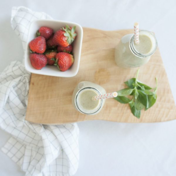 Two Strawberry Basil Smoothies in jars with straws placed on a wooden cutting board with a white bowl of strawberries and basil leaves.