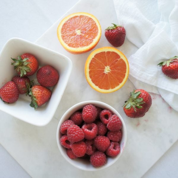 A white bowl of strawberries beside a white bowl of raspberries placed on a white cutting board with a sliced grapefruit.