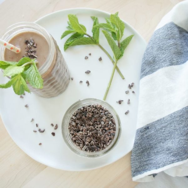 After 8 Chocolate Mint Protein Smoothie poured into a jar class placed on a white serving plate with a jar of chia seeds and a blue and white kitchen towel. Ingredients include almond milk, black beans, cacao nibs, banana, fresh mint, cocoa.
