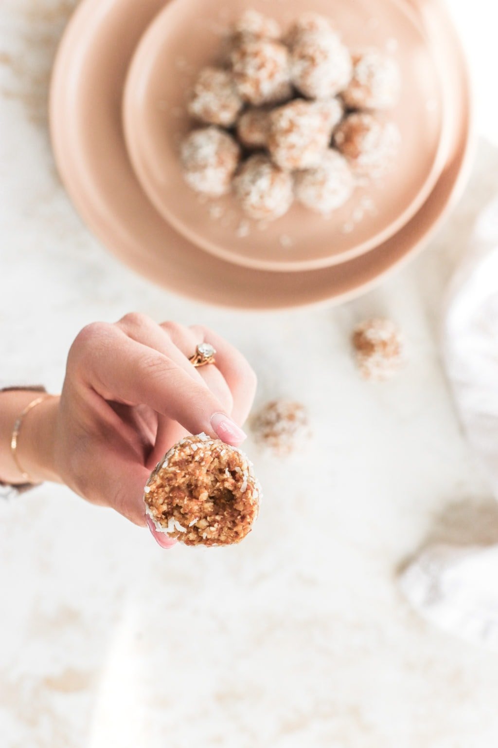 A hand is holding an energy ball with a bite taken out of it. There is a blurred out plate of energy balls in the background.