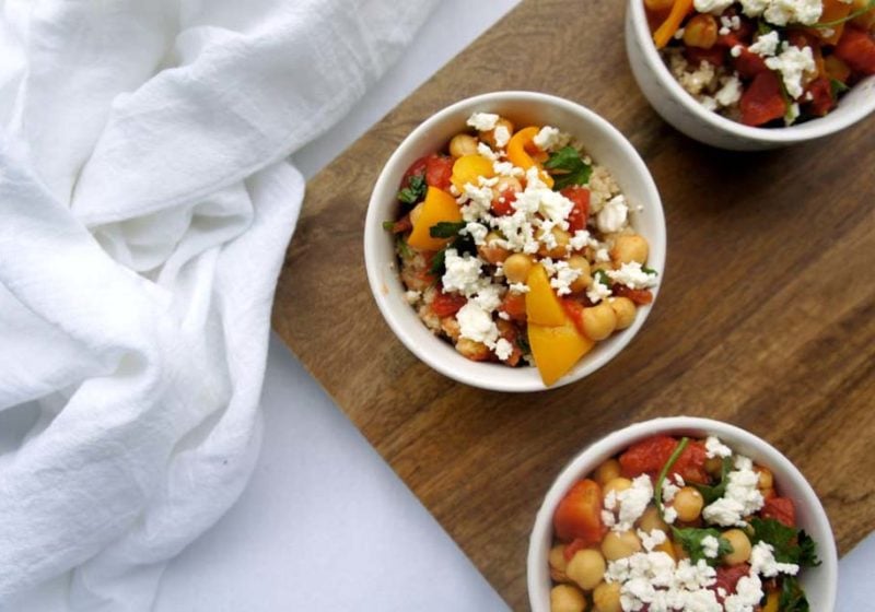 Chickpeas a la Grecque in a white round bowl placed on a wooden cutting board on a white surface with a white kitchen towel. Ingredients include couscous, garlic, bell peppers, chickpeas, tomatoes, vegetable broth, parsley, oregano, feta cheese, lemon wedges.