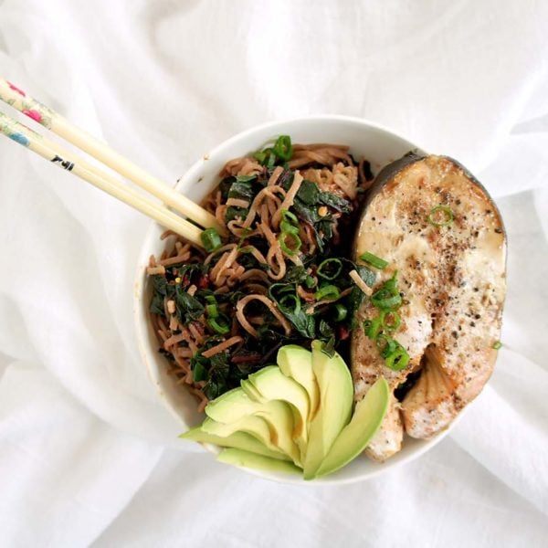 Salmon Soba Noodle Bowl placed in a white round bowl with chopsticks placed on a white sheet. Ingredients include salmon, avocado, soba noodles.
