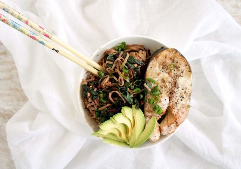 Salmon Soba Noodle Bowl placed in a white round bowl with chopsticks placed on a white sheet. Ingredients include salmon, avocado, soba noodles.