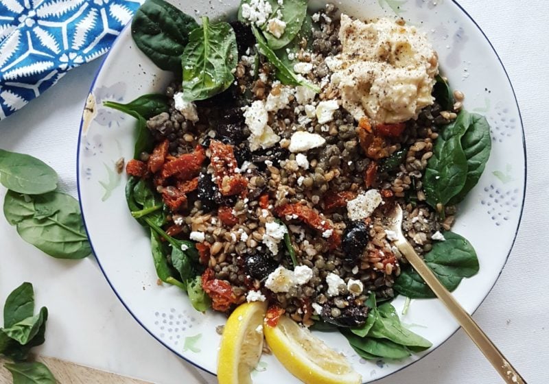 Mediterranean Farro and Green Lentil GRAIN Bowl with Feta, Olives and Sundried Tomatoes placed on a round plate. Ingredients include water, farro, lentils, cheese, black olives, baby spinach, tomatoes, hummus.