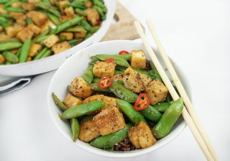 Crispy Black Pepper and Maple Tofu Snap Pea Stir Fry placed in a white round bowl with chopsticks. Ingredients include snap peas, tofu, soy sauce, garlic, ginger, chili, green onions.