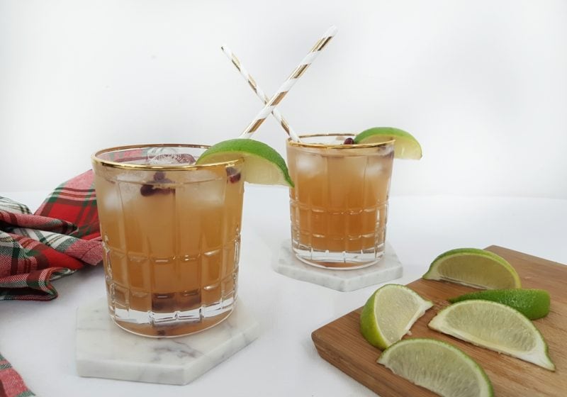 Ginger Pomegranate Kombucha Moscow Mule poured into two glasses with striped straws with a cutting board an lime wedges place beside. Ingredients include ice, vodka, ginger kombucha, pomegranate seeds, lime.