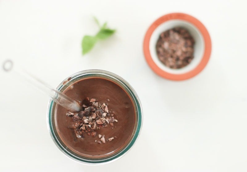An overhead view of a dark chocolate smoothie in a cup with a glass straw and a small bowl of cacao nibs in a bowl beside it. a piece of fresh mint is blurred in the background. Recipe by Lindsay Pleskot Dietitian