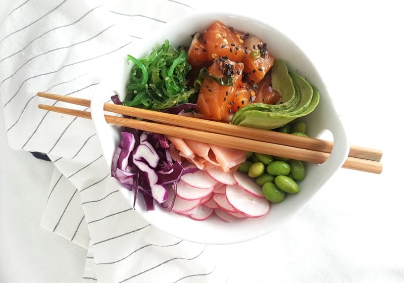A round white bowl of rice, salmon, edamame beans, radishes, cabbage, salad on a white surface with a striped kitchen towel.