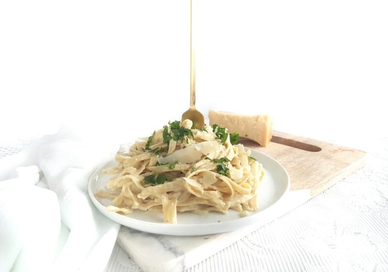 Cauliflower Fettuccini Alfredo on a round white plate placed on a cutting board with a white kitchen towel placed beside. Ingredients include fettuccini noodles, garlic, Parmesan, salt, parsley.