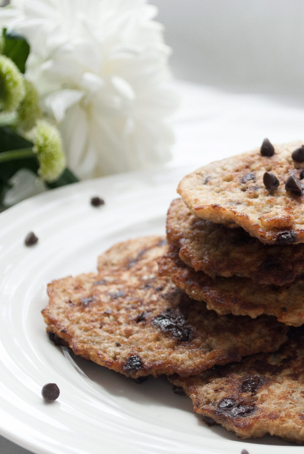 A close up of a stack of 4 banana oat pancakes. The pancakes are topped with chocolate chips. There are white flowers in the background