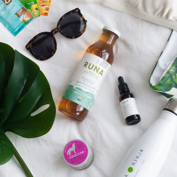 A photo of travel essentials placed on a white sheet. EnerC packets, sunglasses, Runa Tea, skin care, and a water bottle placed by a large green plant.