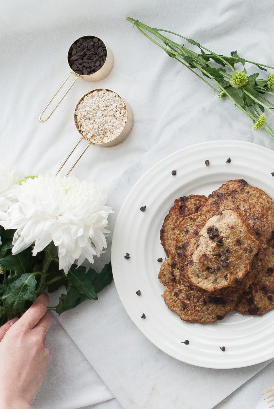 A stack of 5 banana oatmeal pancakes sits on a white plate. There are chocolate chips loosely thrown over the plate. Above the plate is a 1/3 cup measuring cup of chocolate chips and 1 cup measuring cup of rolled oats. There is a hand placing white flowers beside the plate.