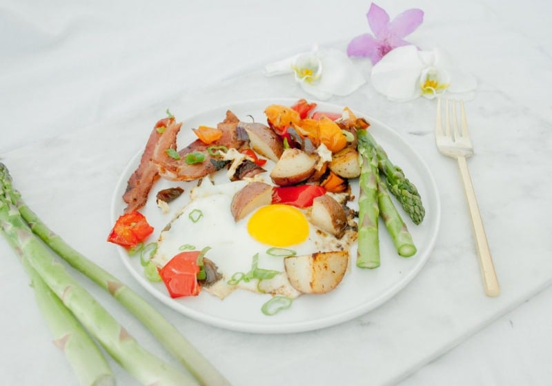 A round white plate placed on a marble cutting board with flowers and asparagus placed around. Ingredients include bacon, eggs, potatoes, bell pepper, onion, avocado oil, asparagus, pepper.