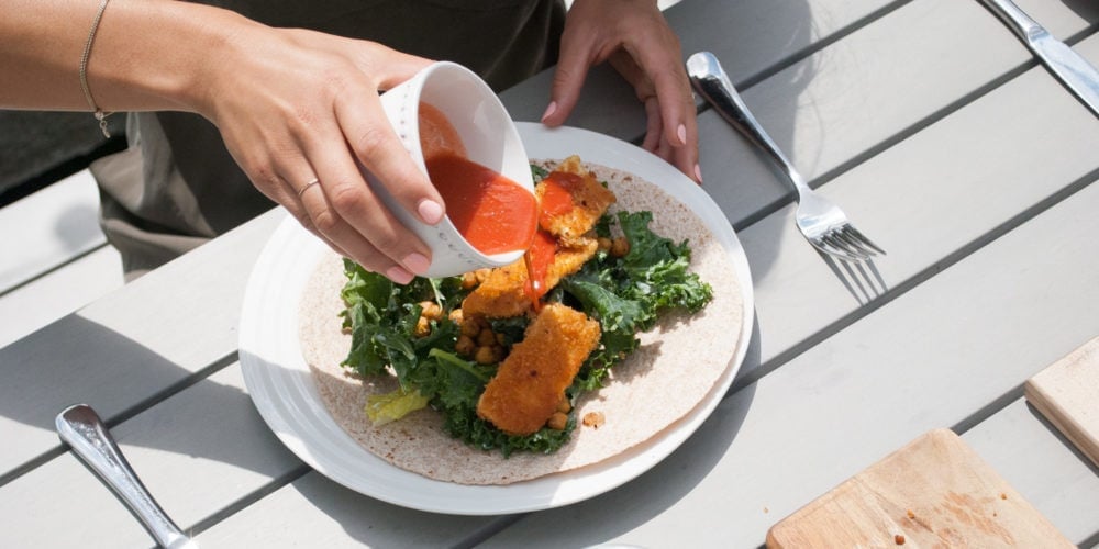 Registered Dietitian Lindsay Pleskot pouring dressing over a Buffalo Tofu Caesar Wrap outside on the patio. Ingredients include whole grain wraps, tofu, breadcrumbs, greens, hot sauce.