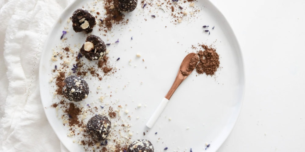 A round white plate topped with Healthy Ferrero Rocher Energy Bites on a white surface with a white kitchen towel. Ingredients include dates, hazelnuts, cacao, sea salt.