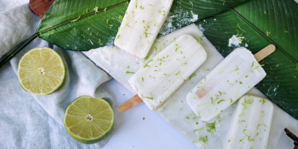 4 Popsicle's scattered over a big green leaf with a cut open lime over a white surface. Ingredients include honey, coconut tequila, triple sec, coconut milk, soda water.
