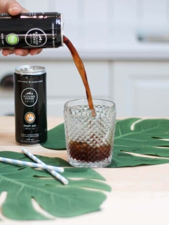 A can of coffee being poured into a drinking glass with two large leaves placed around it.