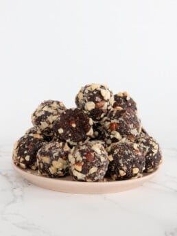 A pile of hazelnut, chocolate energy balls sits on a pink plate on a marble counter top.