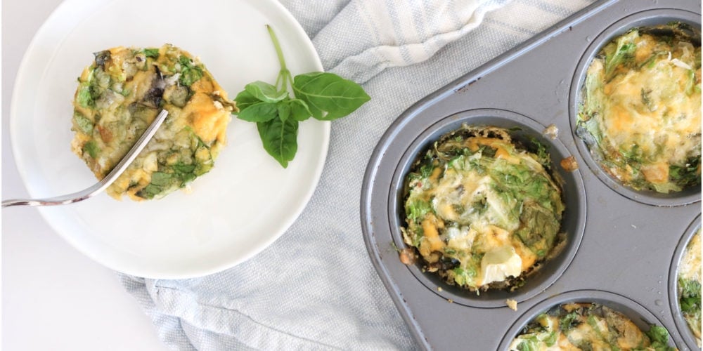 A round white plate with a frittata placed over a light blue kitchen towel and muffin tin filled with frittatas. Ingredients include egg, greens, onion, basil, cheese.