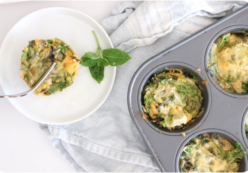 A round white plate with a frittata placed over a light blue kitchen towel and muffin tin filled with frittatas. Ingredients include egg, greens, onion, basil, cheese.
