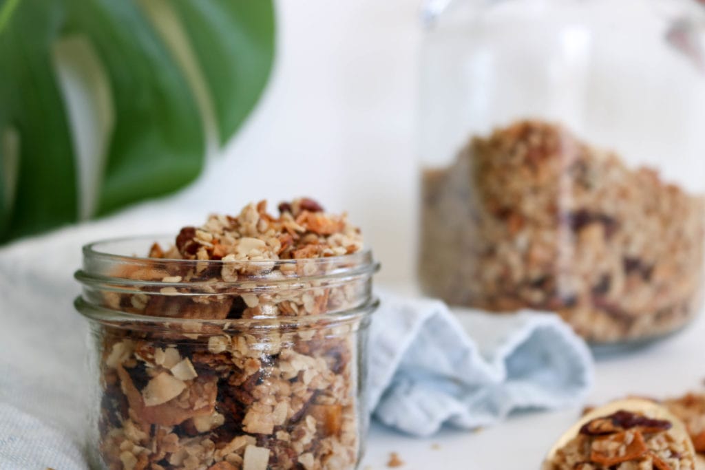 Crunchy & Nutritious Coconut Granola in a mason jar over a white surface with a light blue kitchen towel and greenery. Ingredients include: coconut oil, honey, rolled oats, chia seeds, pecans, walnuts, coconut pieces, dried mango and pineapple.