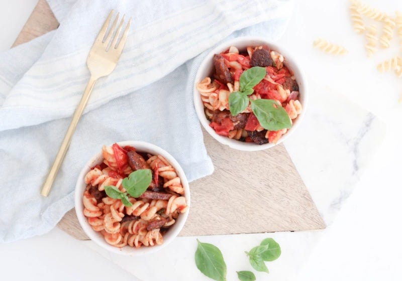 Two white bowls with pasta and a gold fork with a white kitchen towel and basil leaves beside them. Ingredients include penne noodles, tempeh, fennel, onion, garlic, chili.